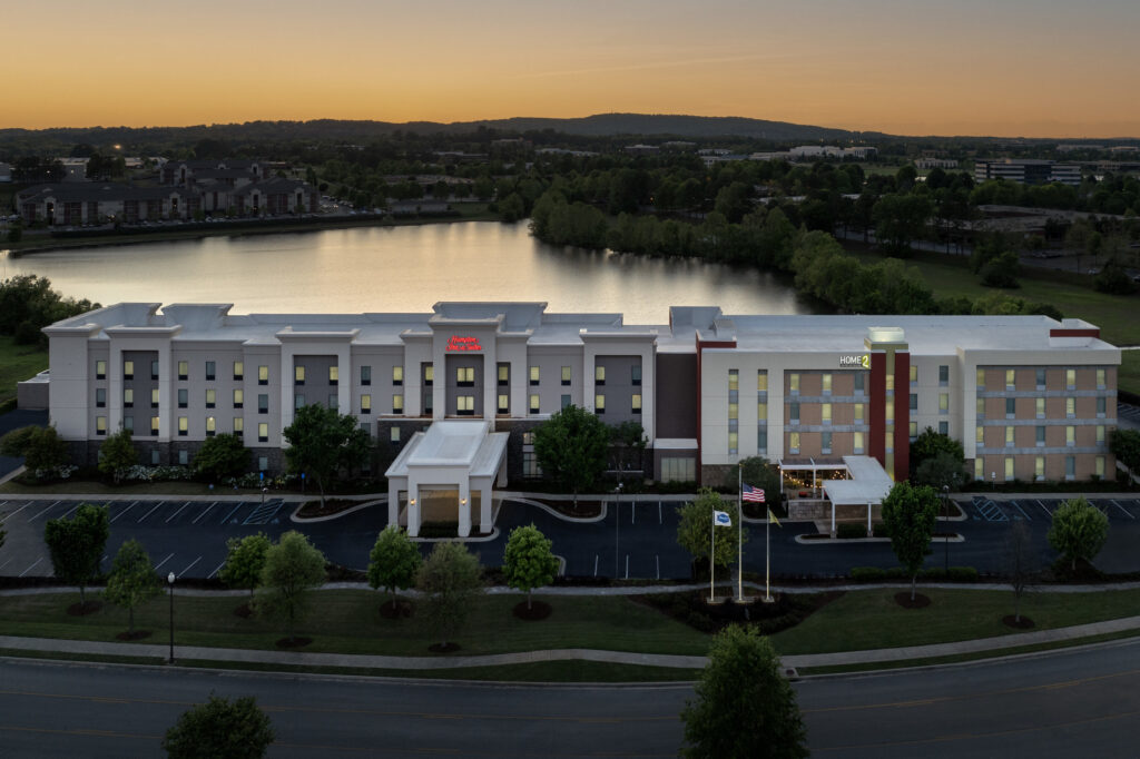 Drone photo of exterior front of Home2 Suites and Hampton Inn and Suites Huntsville Research Park at dusk with lake visible behind it.